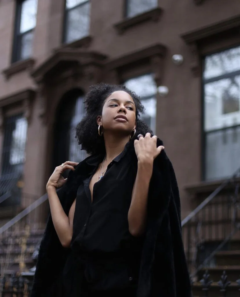 A young woman with curly hair and hoop earrings standing outside in front of brownstone buildings, wearing a black outfit and posing confidently.