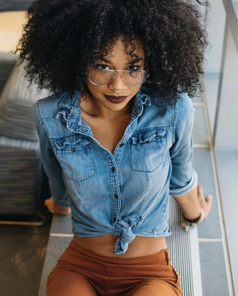 A woman with curly black hair, wearing glasses and dark lipstick, sitting on a bench indoors, looking up at the camera. She is wearing a tied-up denim shirt and rust-colored pants.