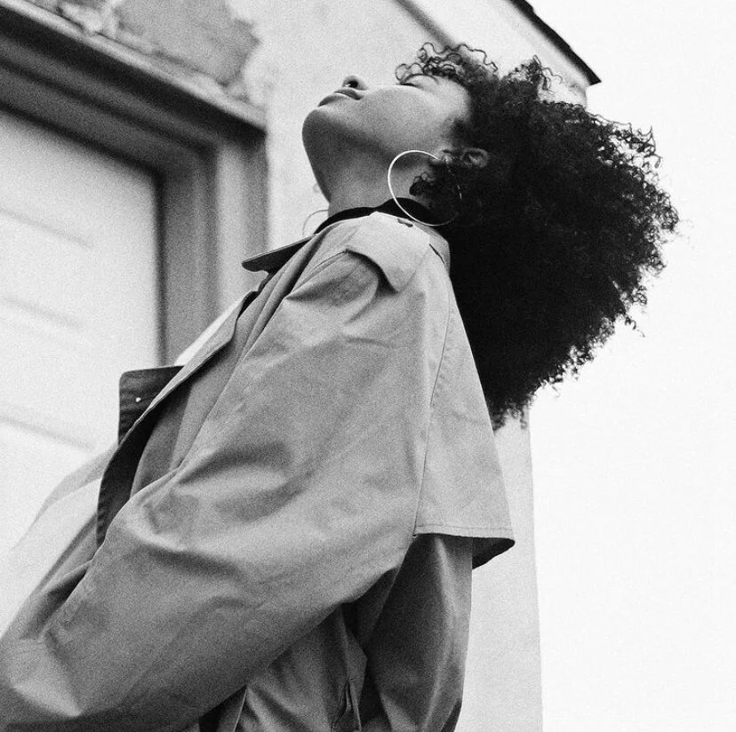 A woman with curly hair and large hoop earrings tilts her head back, wearing a trench coat outdoors in black and white.