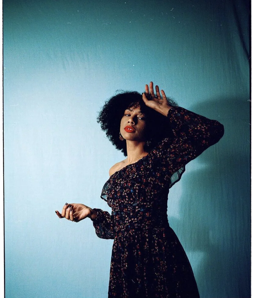 A woman with curly hair and red lipstick poses against a gradient blue background, wearing a dark floral off-shoulder dress.