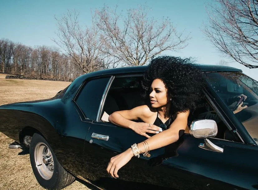 A woman with curly hair leaning out of a black vintage car parked on a grassy field with leafless trees and a clear blue sky in the background.