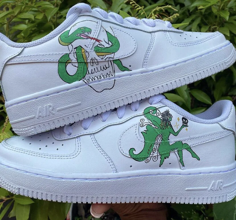 Pair of white Nike Air Force 1 sneakers with custom hand-drawn artwork featuring a green snake intertwined with a skull and a green female figure with curly hair holding a staff with a skull, set against a background of green foliage.
