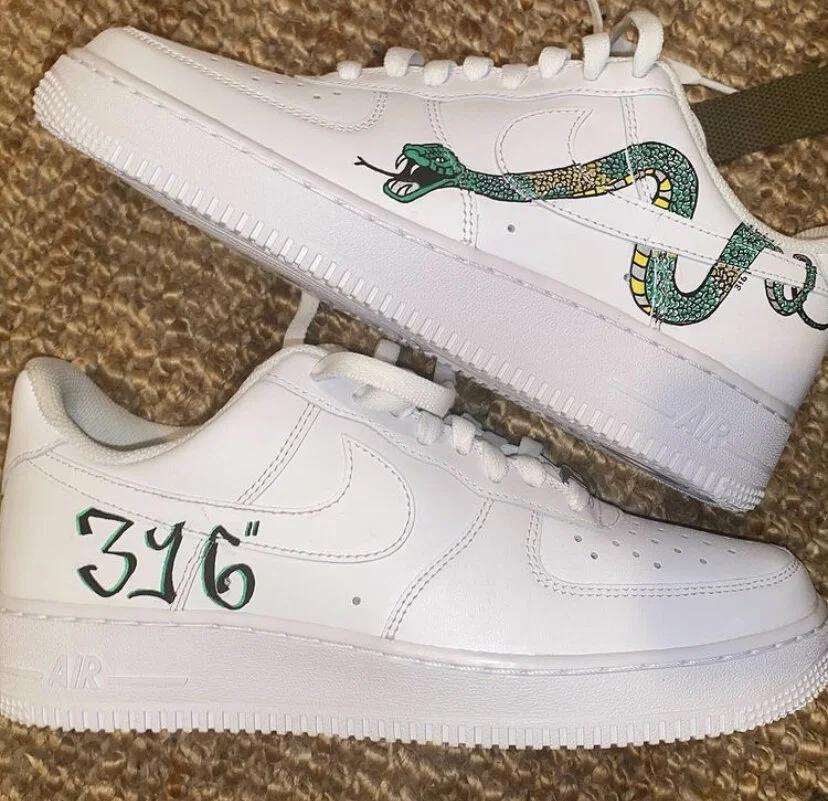 Pair of white Nike Air Force 1 sneakers on a carpeted surface, one decorated with a green snake graphic on the side, and the other with the number "316" written on the side.