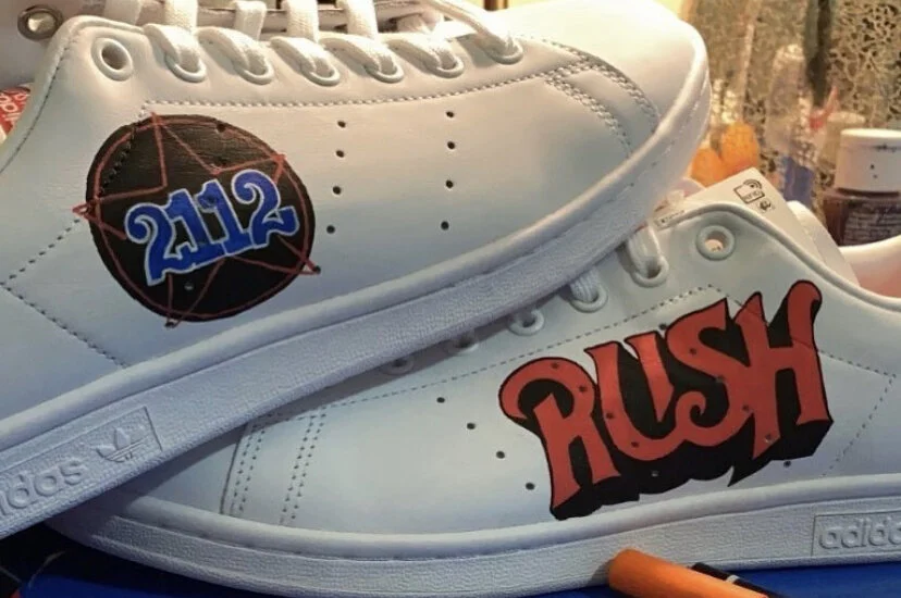 White sneakers with custom designs, including a black circle with a red network and blue "2112" on the left shoe, and the word "RUSH" in red and black on the right shoe.