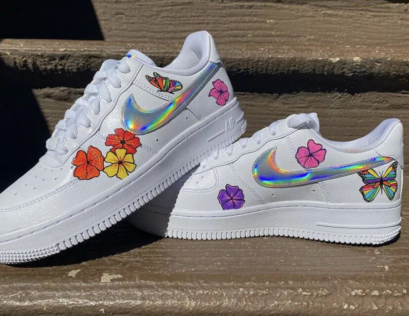 Pair of white sneakers decorated with colorful flower and butterfly stickers, and holographic Nike swoosh logos.