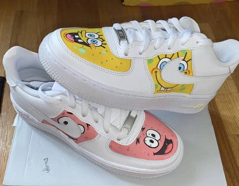 Pair of white sneakers featuring cartoon characters from SpongeBob SquarePants on the toes, with one showing SpongeBob's face and the other showing Patrick Star's face, placed on a white box on a wooden surface.