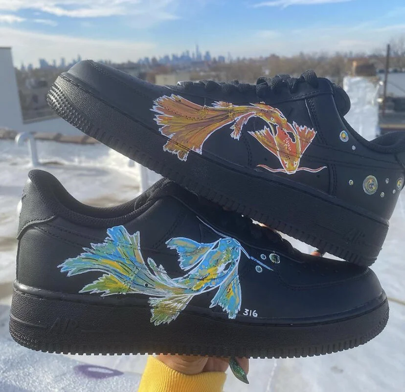 Two black sneakers with painted fish designs, one in orange and the other in blue, outdoors on a snowy rooftop with a city skyline in the background.