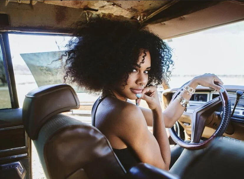 A young woman with curly hair sits in the driver's seat of a vintage vehicle, looking over her shoulder at the camera with her hand resting on her chin, outside scenery visible through the windshield.