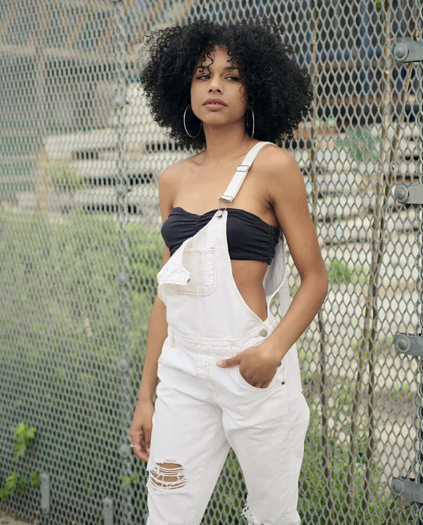 A young woman with curly black hair and hoop earrings posing outdoors in front of a metal fence, wearing a black tube top, white ripped overalls, and standing with one hand in her pocket.