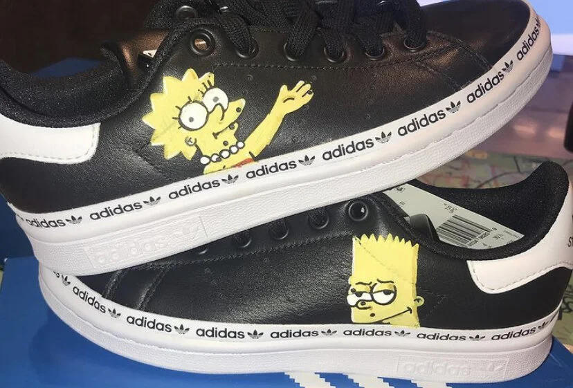 Pair of black Adidas sneakers featuring yellow cartoon characters, Lisa Simpson and Maggie Simpson, printed on the sides.