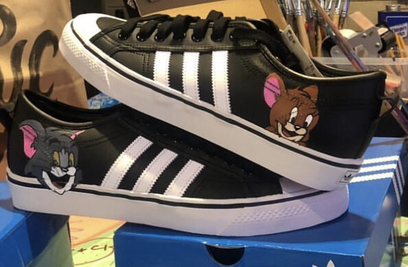 Black sneakers with white stripes and cartoon characters of Tom and Jerry on the sides, placed on a blue shoebox.