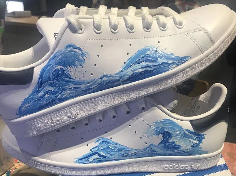 A pair of white Adidas sneakers with blue painted waves on the sides.
