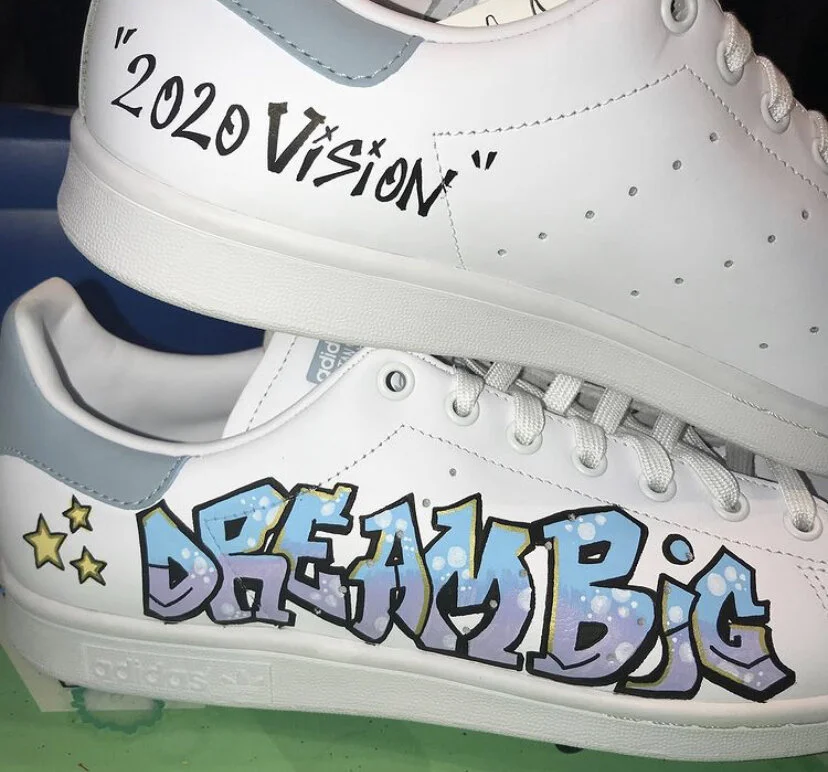 White sneakers with graffiti-style text, one shoe says "2020 VISION" and the other says "DREAM BIG" with colorful dots and stars.