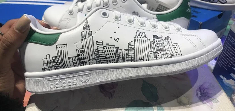 White Adidas sneaker with a black cityscape illustration and green accents on the heel.