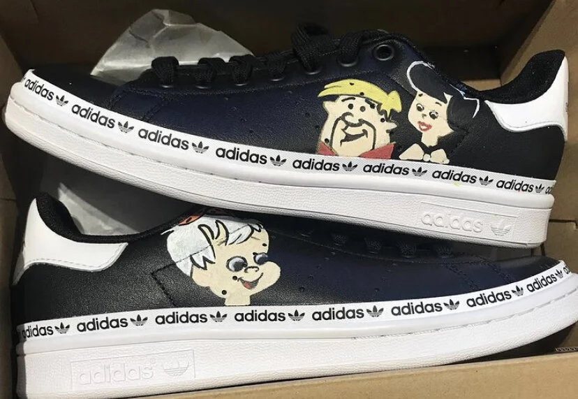Pair of black and white Adidas sneakers with cartoon characters from The Flintstones painted on the sides.