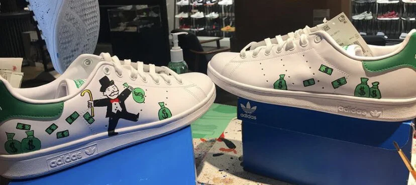 White sneakers with green accents and doodles of money bags, dollar bills, and a cartoon man holding a money bag and a cane, displayed on blue boxes in a store.