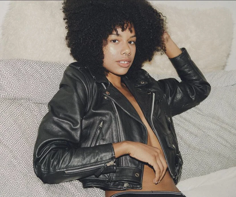 A woman with curly black hair is reclining on a bed, wearing a black leather jacket with no shirt underneath, and is looking at the camera.