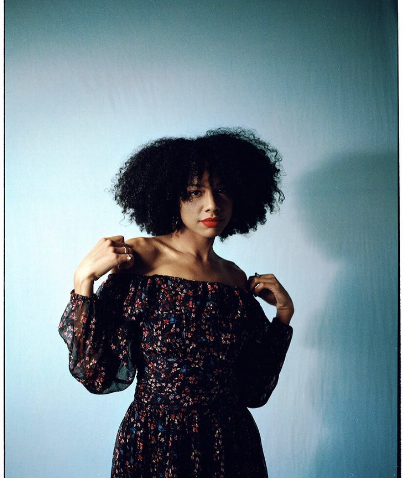 A woman with curly black hair, wearing a black off-the-shoulder dress with red, blue, and white floral pattern, posing against a plain light-colored background.