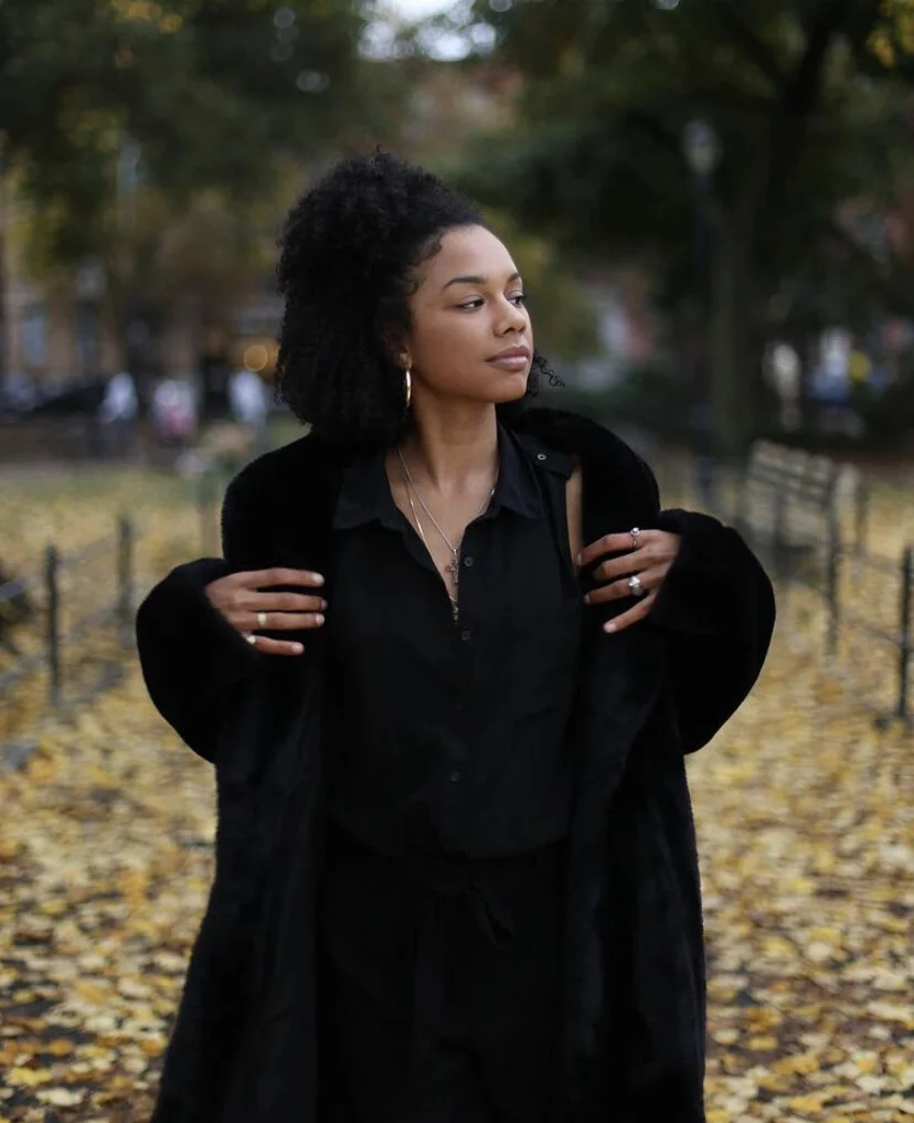 A woman with curly black hair, dressed in black, standing outdoors on a fall day with yellow leaves on the ground and trees in the background.