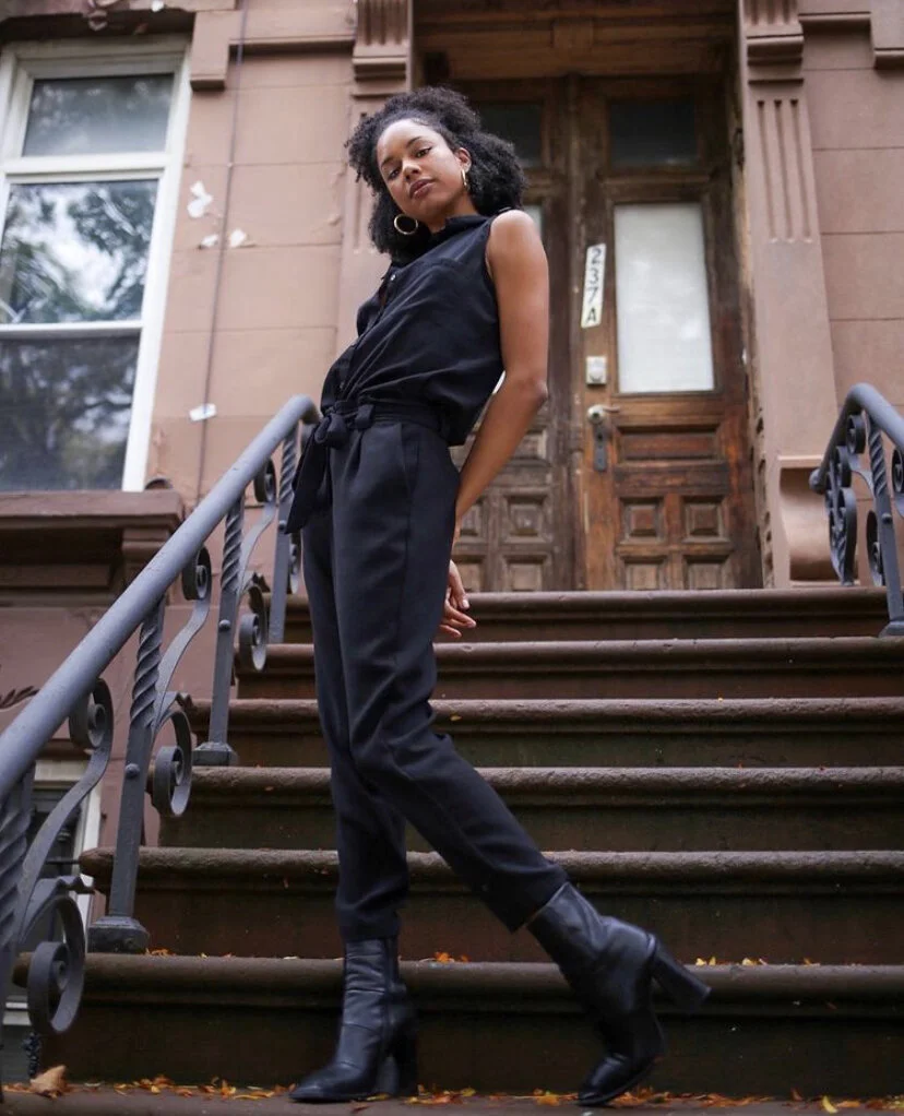 A young woman with curly black hair stands on the steps of a brownstone house, wearing a black sleeveless jumpsuit and black heeled boots, posing confidently.