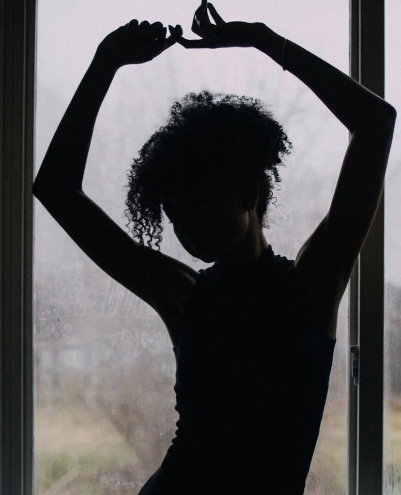 Silhouette of a woman with curly hair standing in front of a rainy window, stretching her arms above her head.
