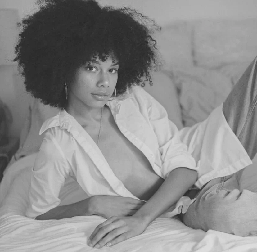 Black and white photo of a woman with curly hair, sitting on a bed, wearing an open shirt, looking at the camera.
