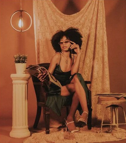 A woman with curly hair sitting on a vintage chair, holding a phone to her ear and a magazine. She is wearing a black dress and white high heels, in a warmly lit room with orange walls, a round hanging light, a speaker on a small table, and a curtain