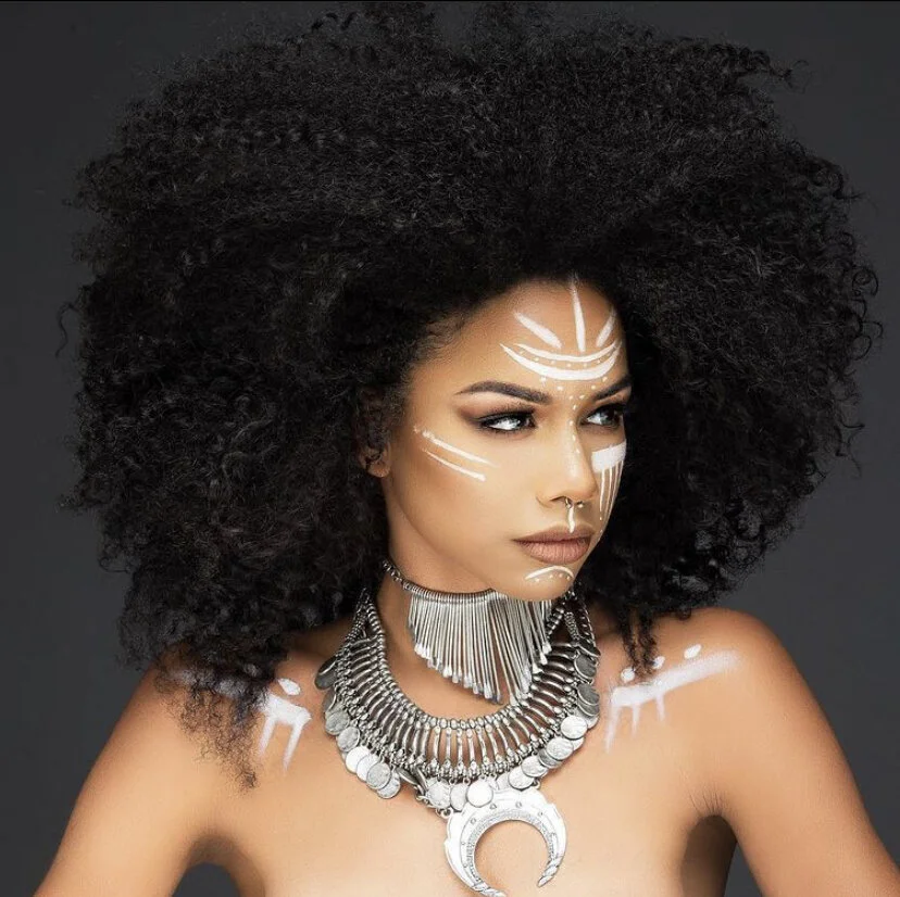 A woman with voluminous curly hair and bold tribal face paint, wearing multiple layered silver necklaces and a large crescent-shaped pendant, against a dark background.