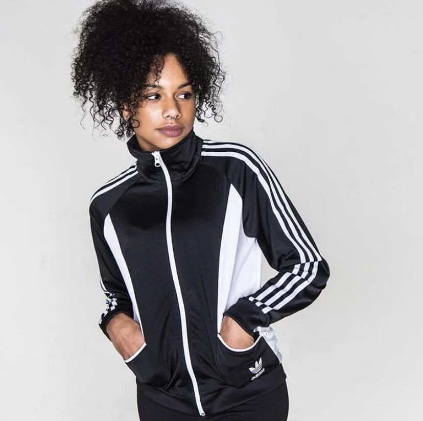 A woman with curly black hair, wearing a black and white Adidas track jacket with her hands in the pockets, standing against a plain gray background.
