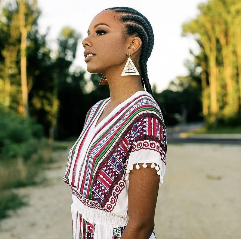 Young woman with braided hair wearing a colorful embroidered dress and large geometric earrings outdoors