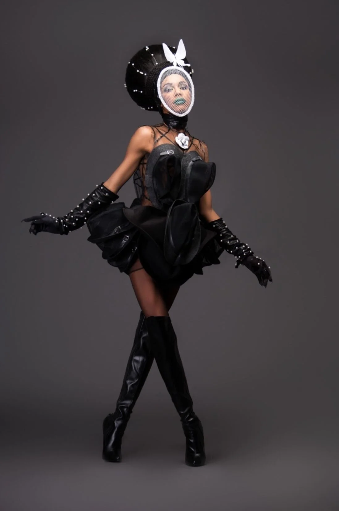 A person dressed in black with avant-garde fashion makeup and accessories, including a large headpiece, sheer top, and thigh-high boots, posing against a dark gray background.
