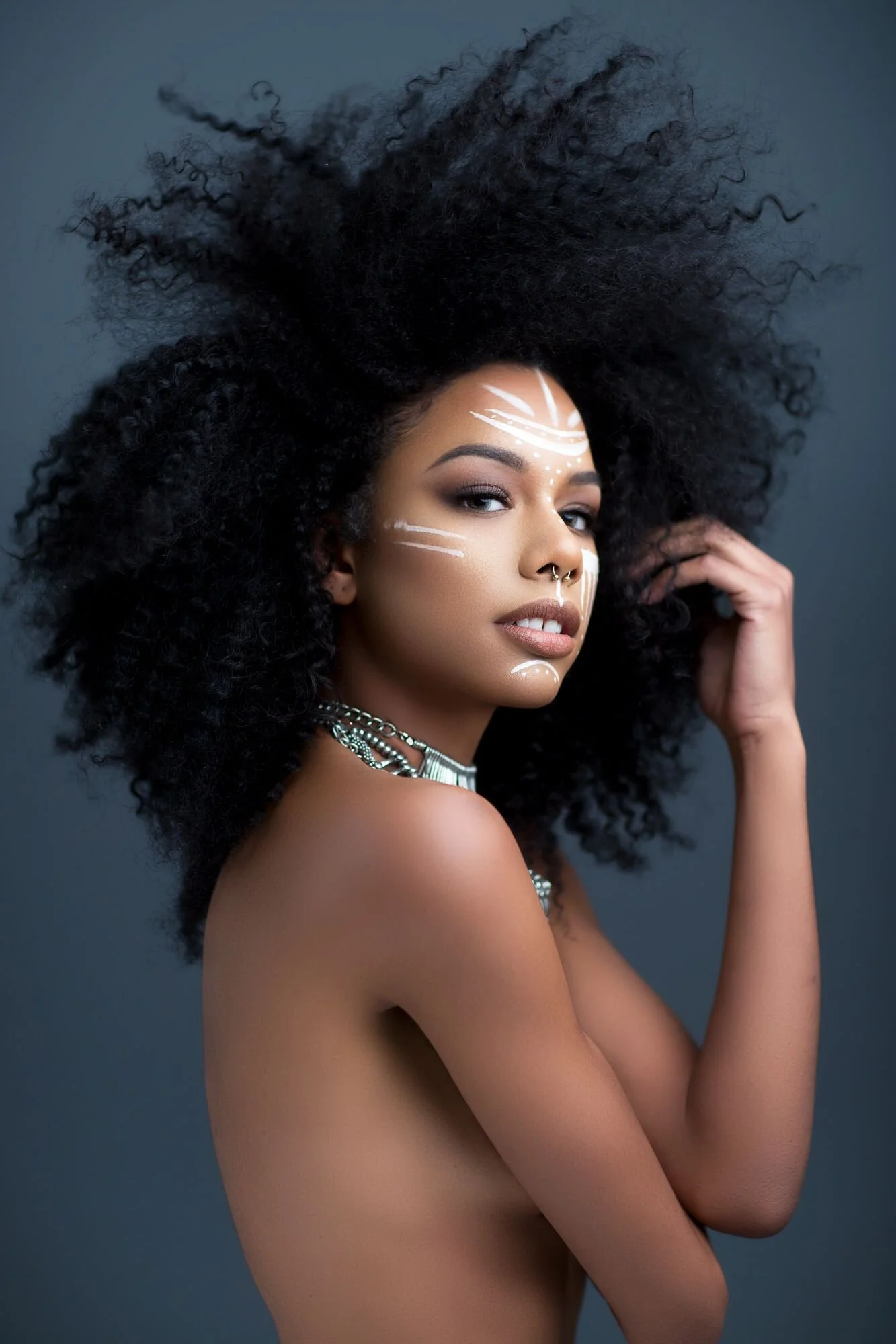 Portrait of a woman with natural curly hair, adorned with white tribal-style face paint, wearing silver jewelry, with a gray background.
