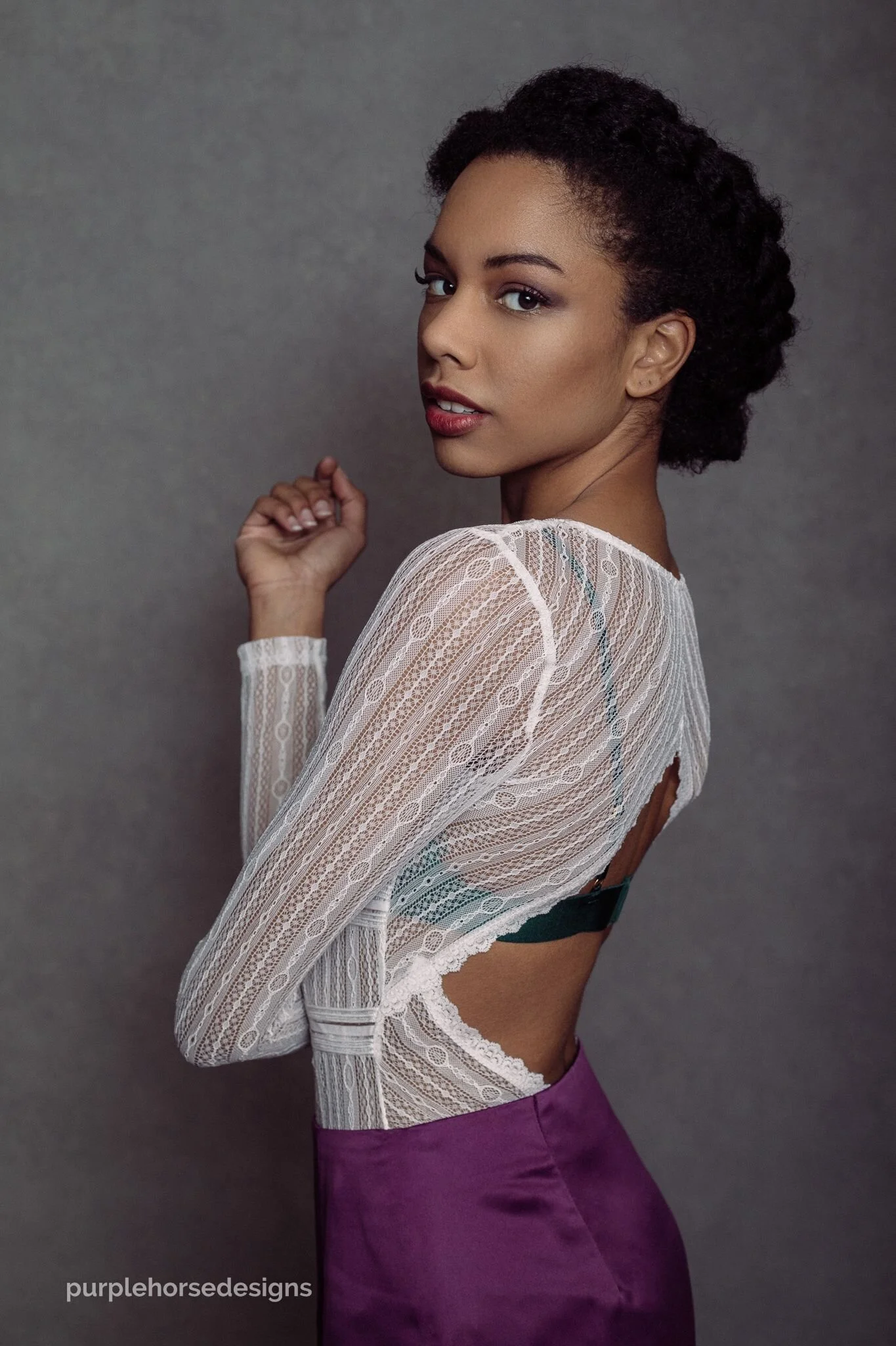 A woman with dark curly hair wearing a sheer white lace top and purple pants standing against a gray background.