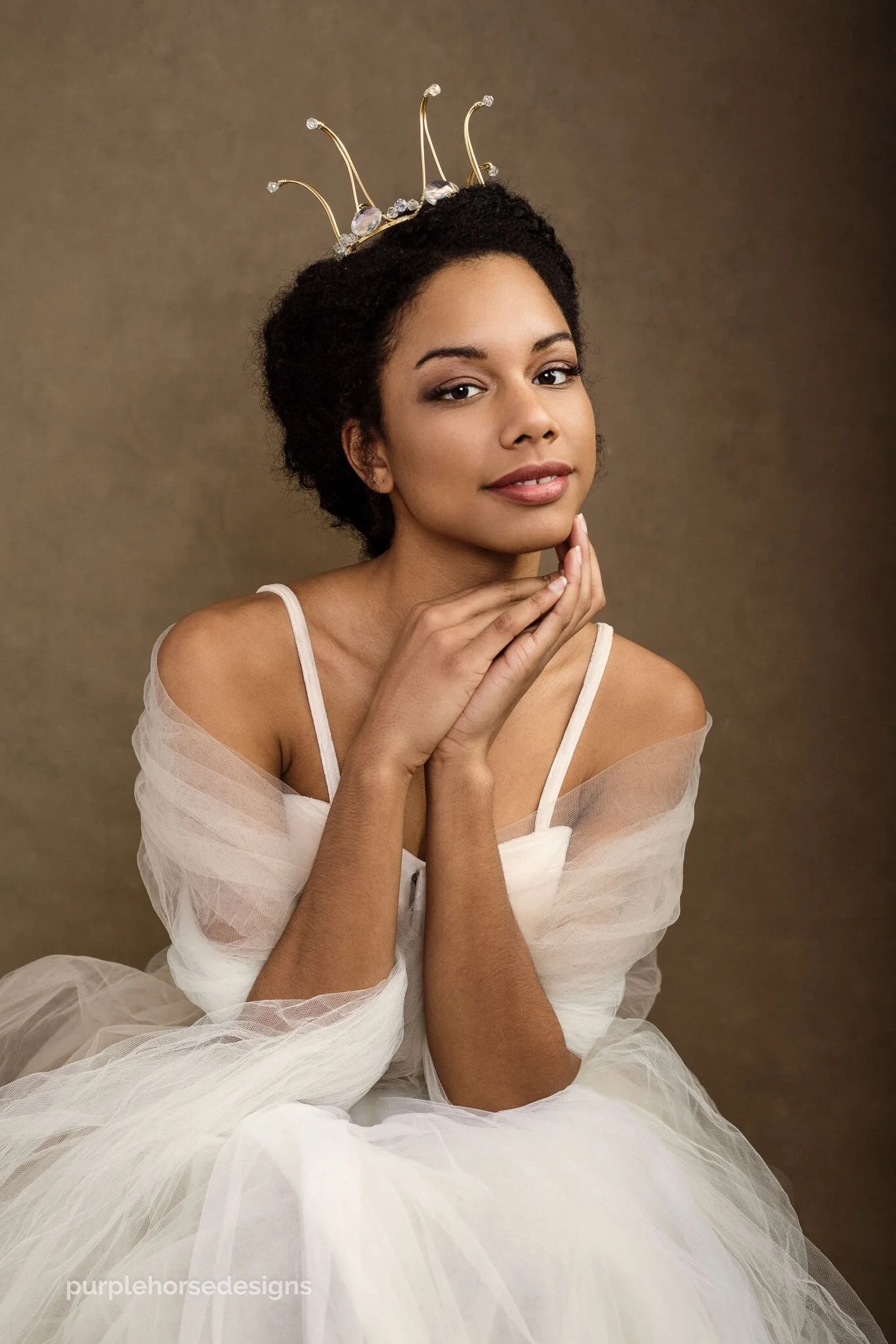 A young woman with dark hair styled in an updo, wearing makeup, a white off-shoulder dress with tulle fabric, and a rhinestone tiara. She has a gentle smile, with her hands resting under her chin, against a plain, warm-toned background.