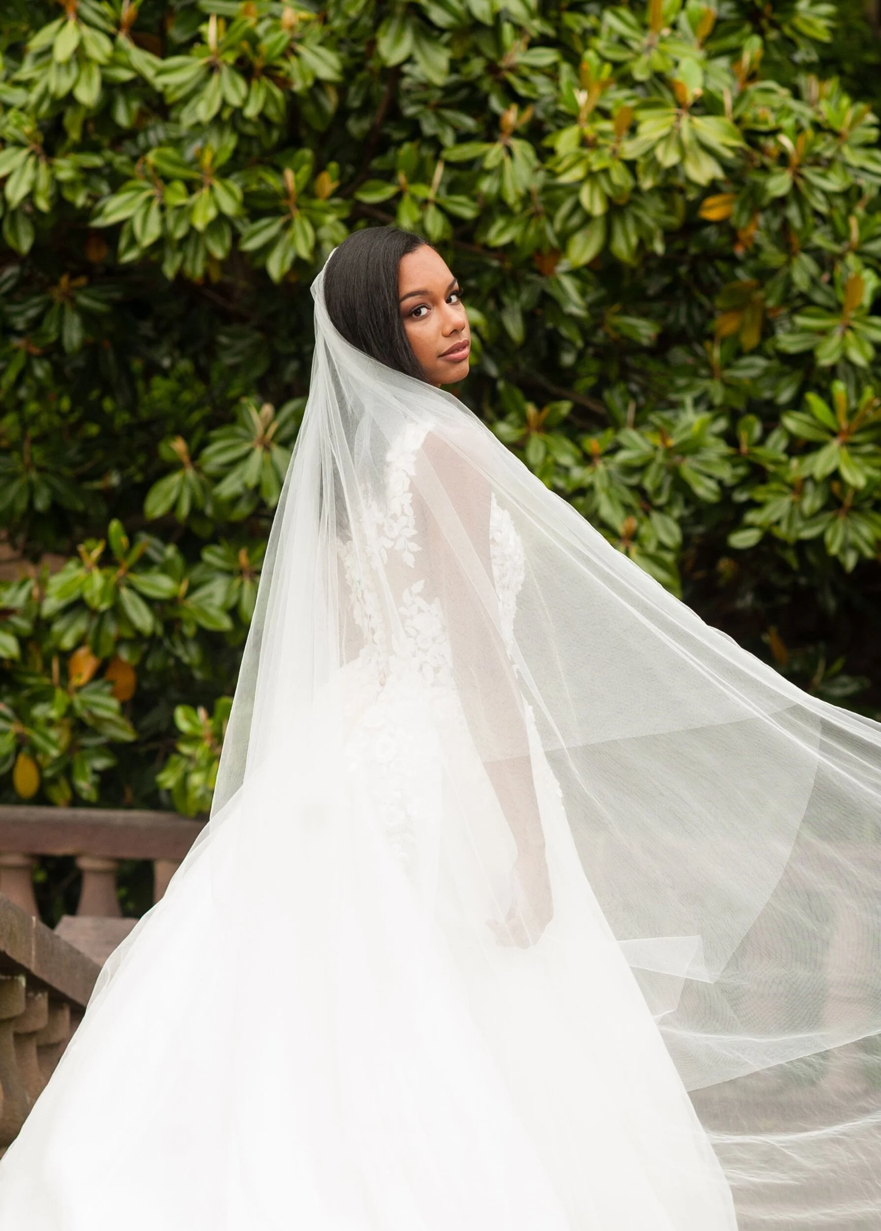 A woman in a white wedding dress with a veil standing outdoors in front of green leafy bushes.