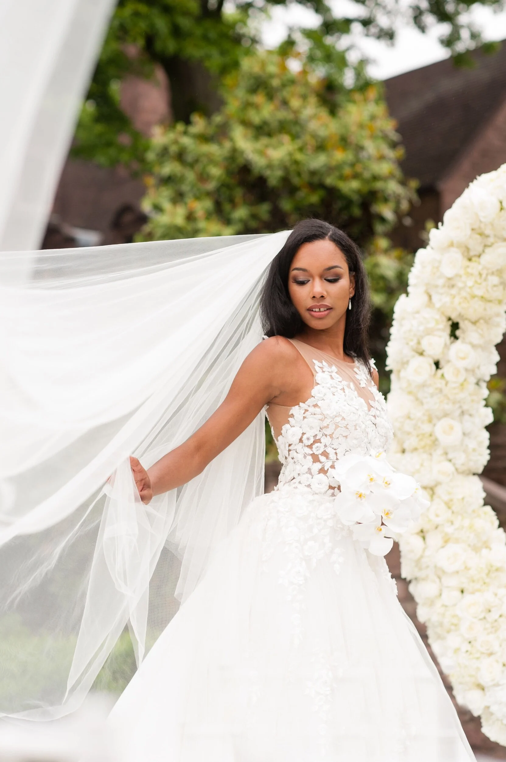 A bride wearing a white wedding dress with floral lace details and a long, flowing veil stands outdoors near a large floral arrangement of white roses and orchids, with trees and a building in the background.