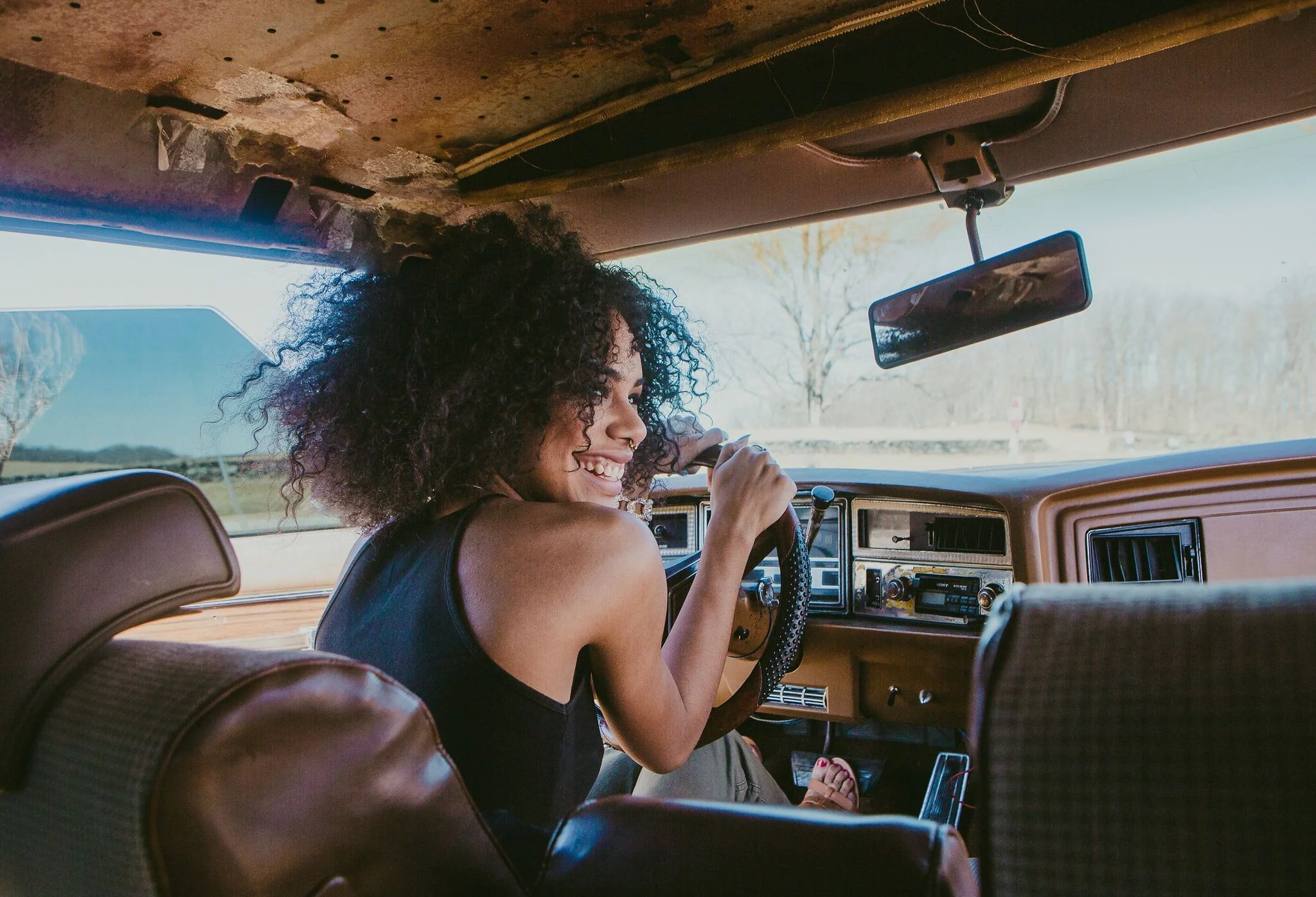 Young woman with curly hair smiling as she drives an orange vintage car on a sunny day