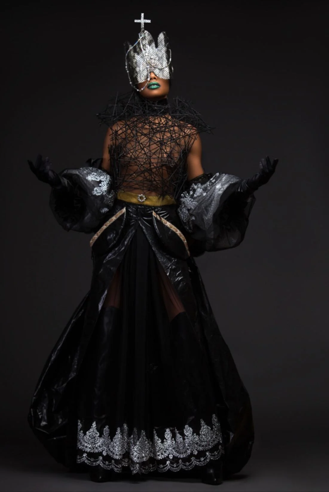Person wearing an elaborate black and silver fashion ensemble with mesh and embroidered fabric, metallic face covering, and statement jewelry, standing against a dark background.