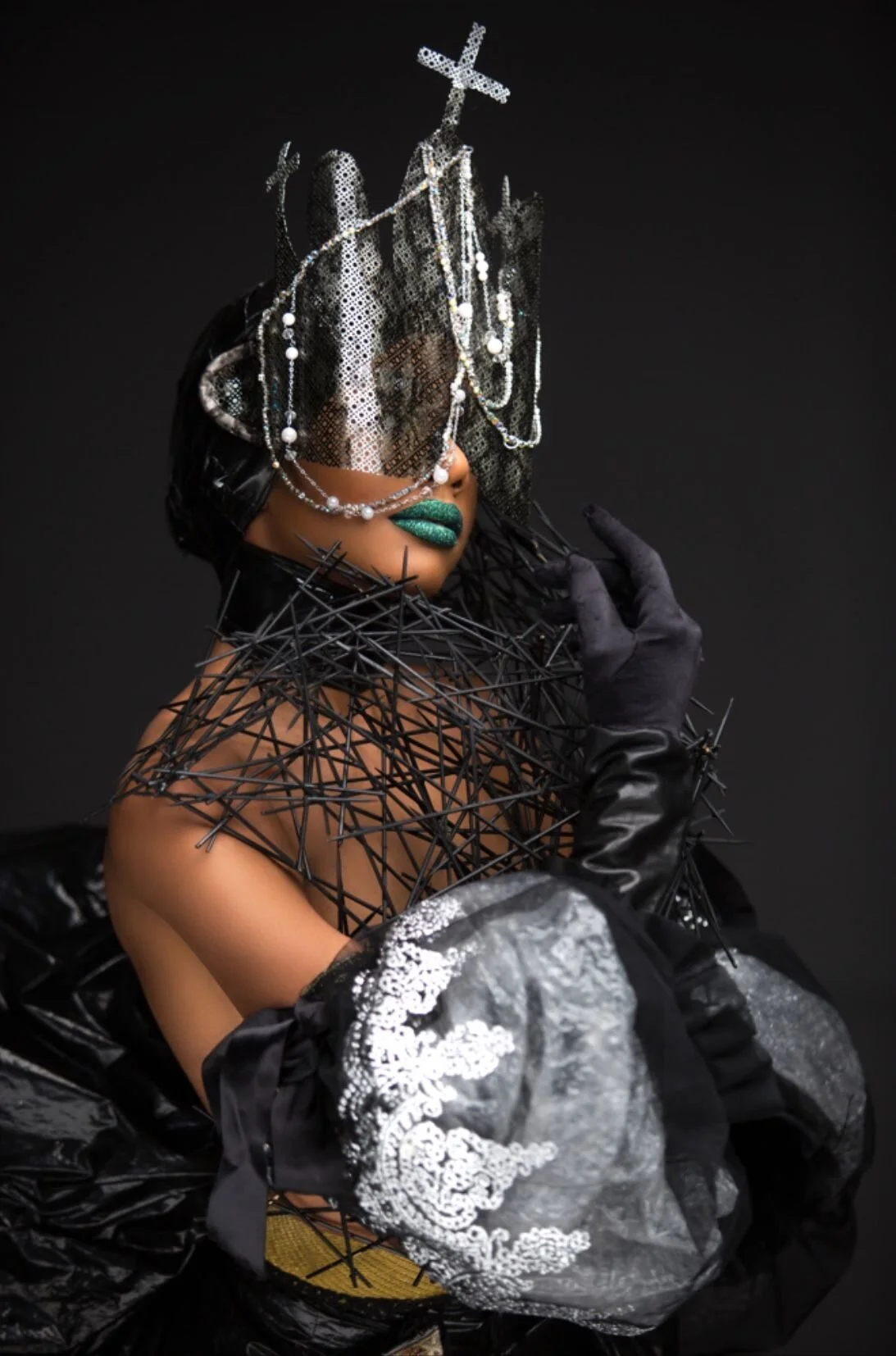 A woman with artistic makeup wearing a headpiece with crosses and pearls, black gloves, and a dress with black and gray fabric and patterns. She has green lipstick and is posed against a black background.