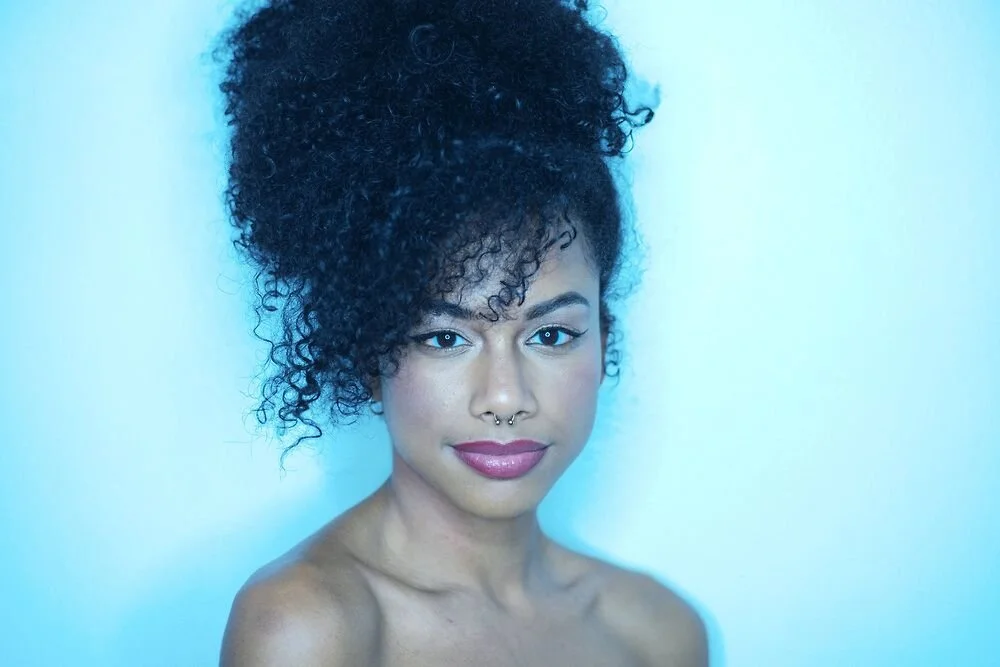 Portrait of a woman with curly dark hair styled in an updo, wearing makeup, and a septum piercing, against a light blue background.