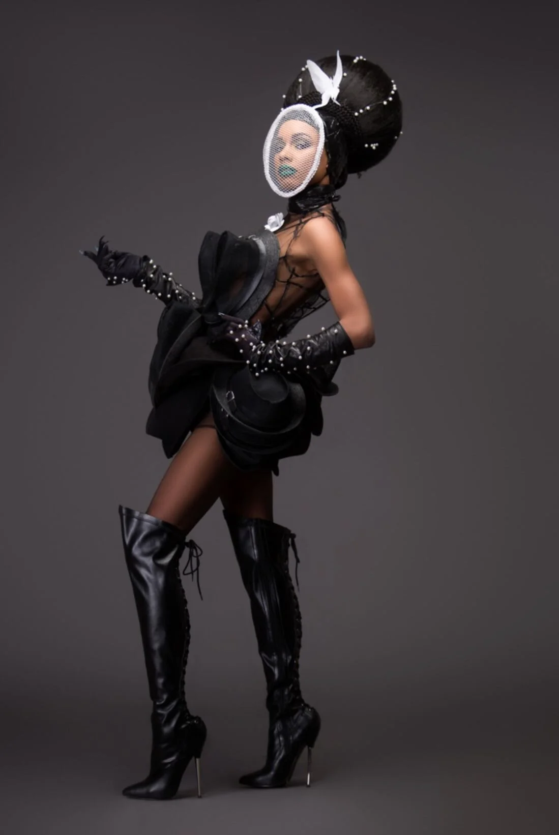 A fashion model wearing black thigh-high boots, a black sheer dress with ruffled details, gloves with decorative pearls, and an elaborate headpiece with a large beaded hat, birdcage veil, and feather accents, posing against a plain dark background.