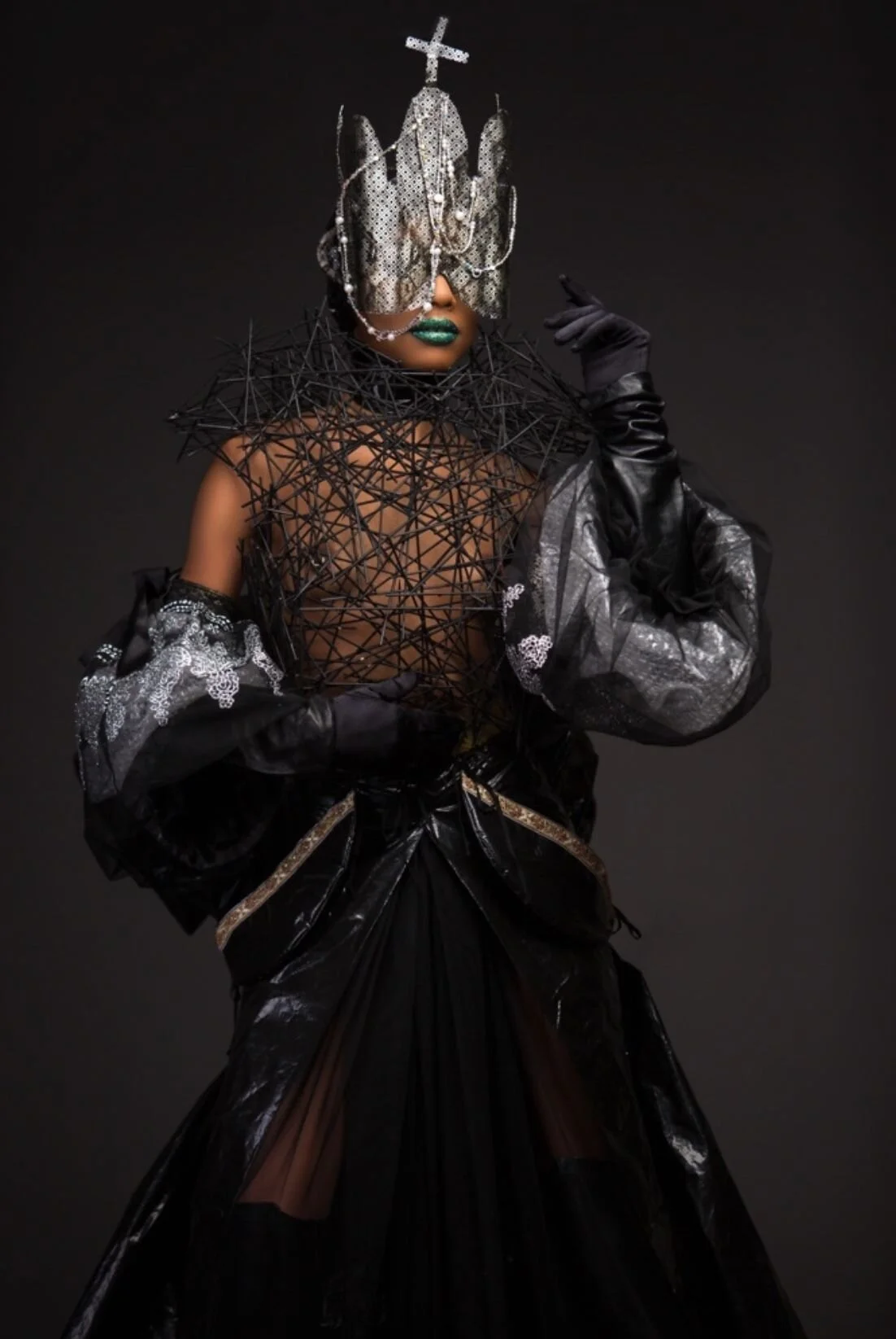 A person wearing an elaborate, avant-garde fashion outfit with a metallic mask and headpiece, black gloves, and a dress with intricate designs against a black background.