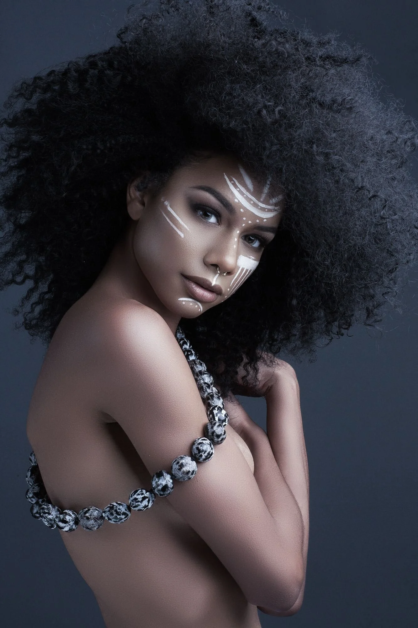 A woman with dark skin and large, curly black hair styled in an Afro, with white tribal face paint, is posed with her arm across her chest, wearing a chunky black and white beaded necklace, against a dark background.