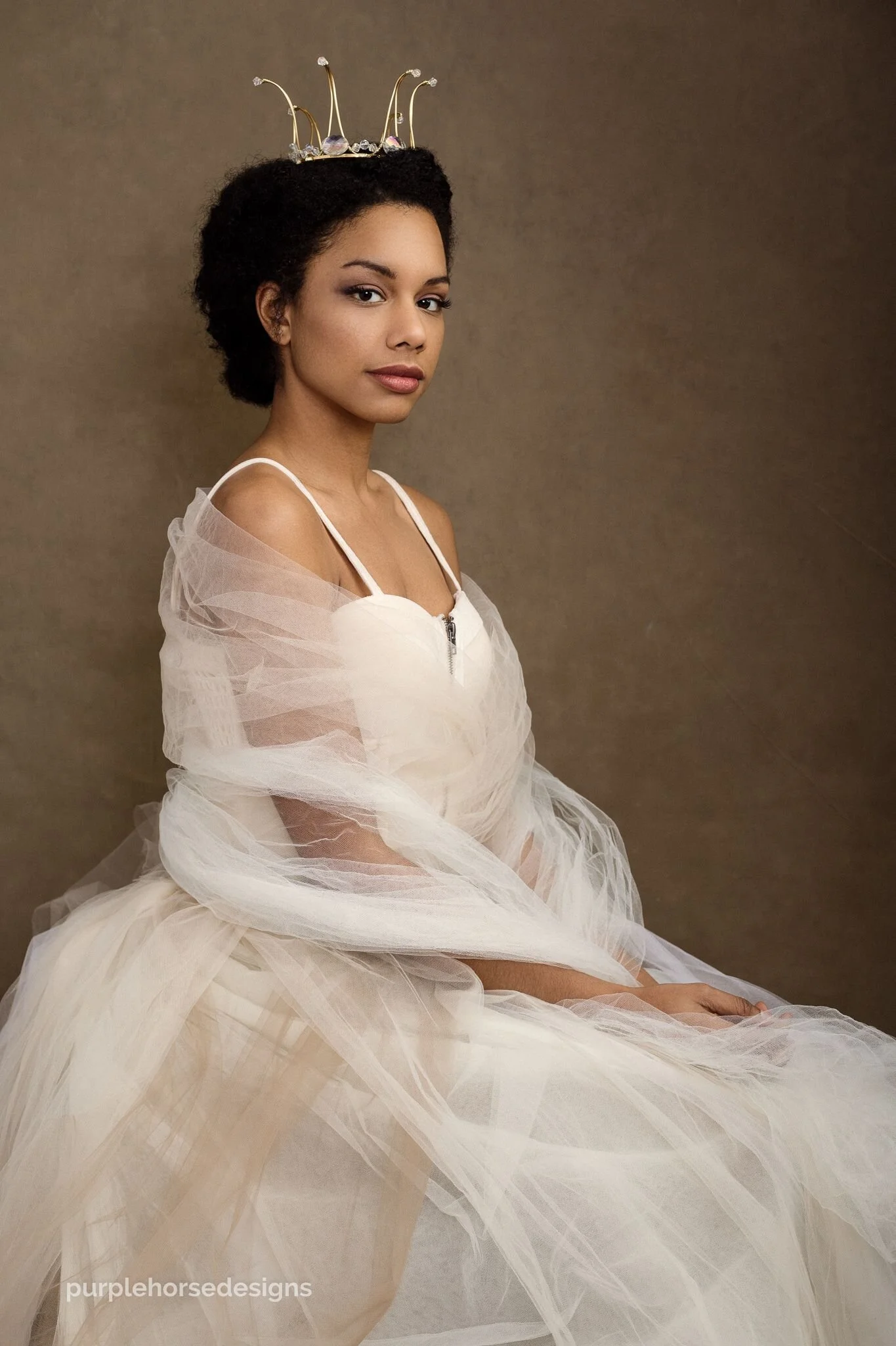 A woman with dark curly hair wearing a crown with wires and decorative crystals, dressed in a beige tulle gown with off-shoulder straps, sitting against a plain background.