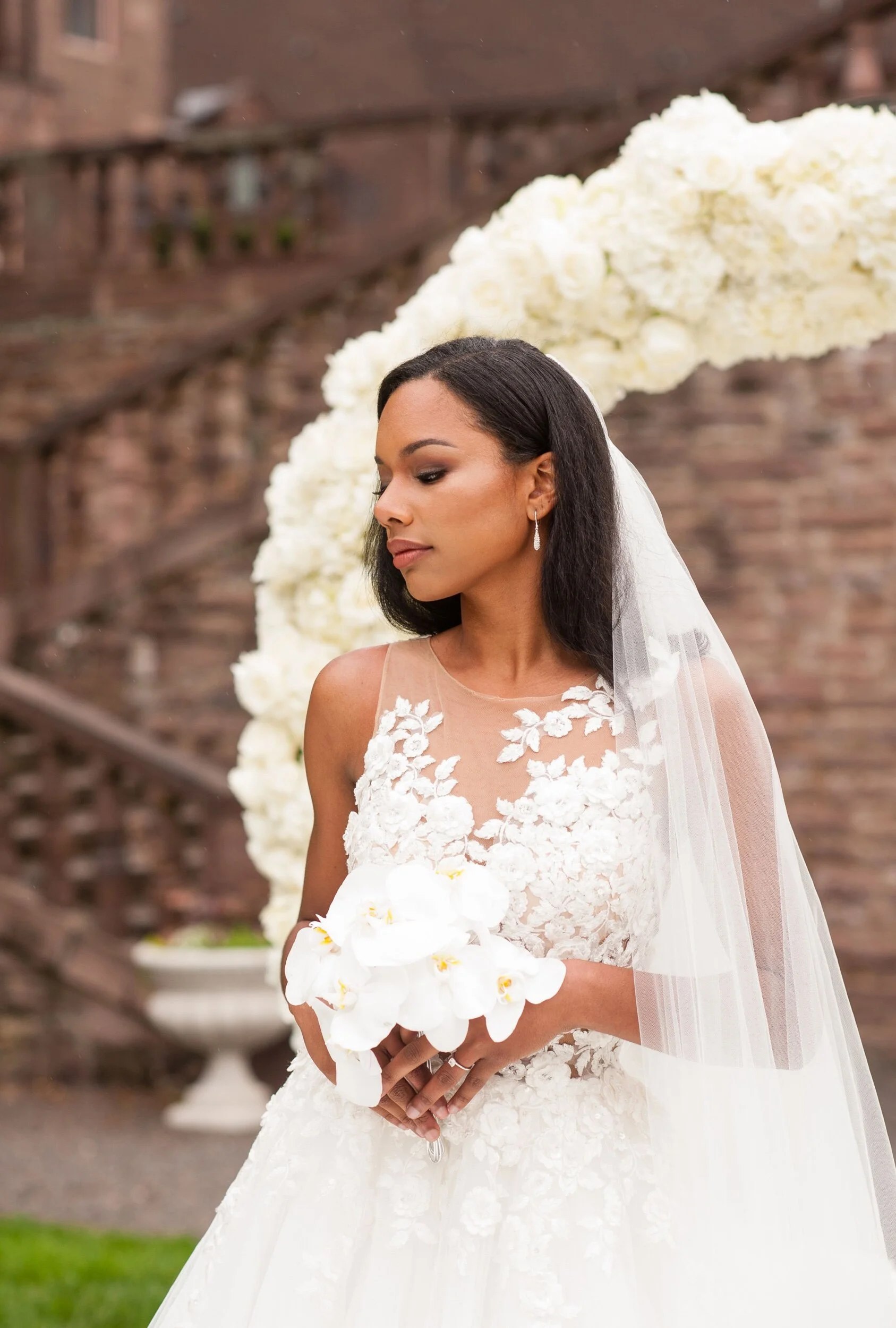 A bride standing outdoors with a large floral arrangement behind her, wearing a white wedding dress with floral lace details and holding a bouquet of white orchids.