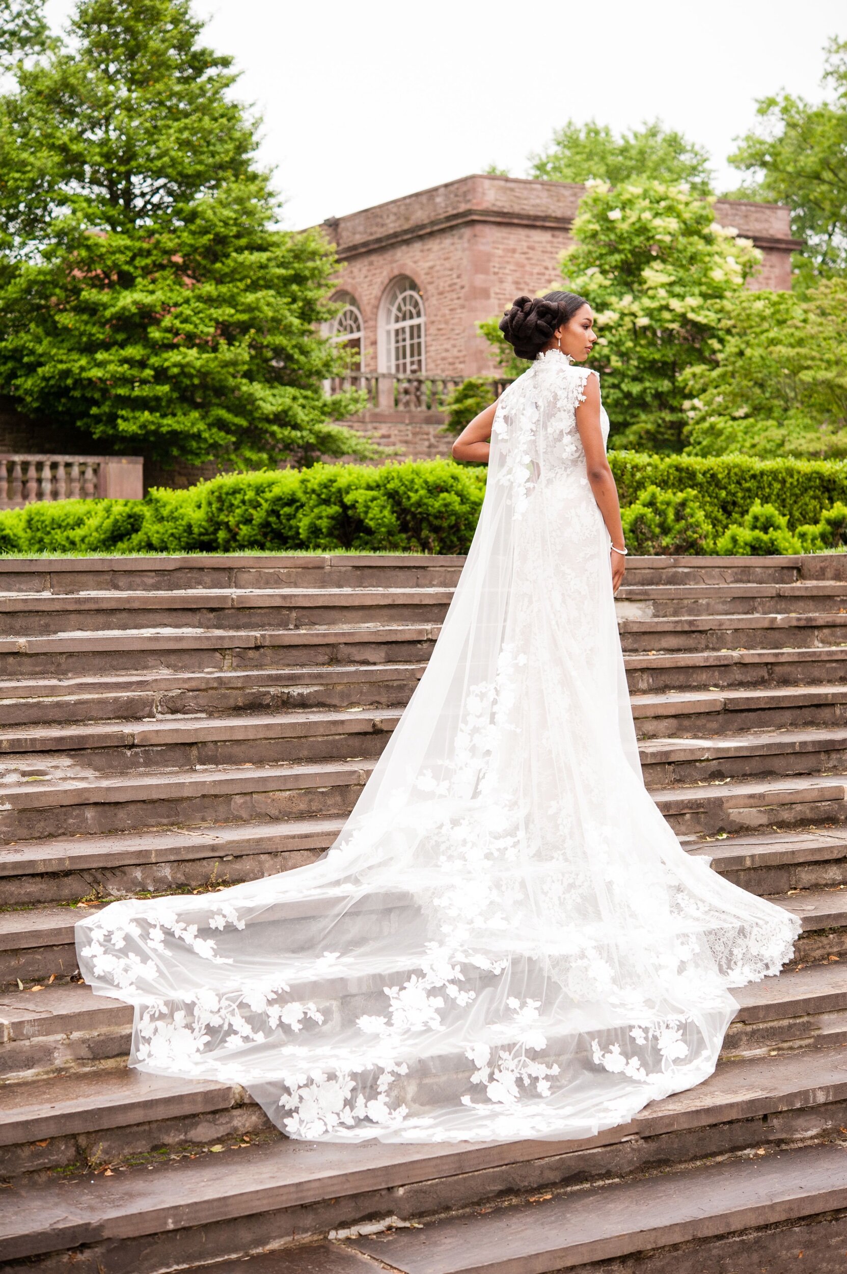 A woman in a white lace wedding dress standing on stone steps outdoors with green trees and an old brick building in the background.