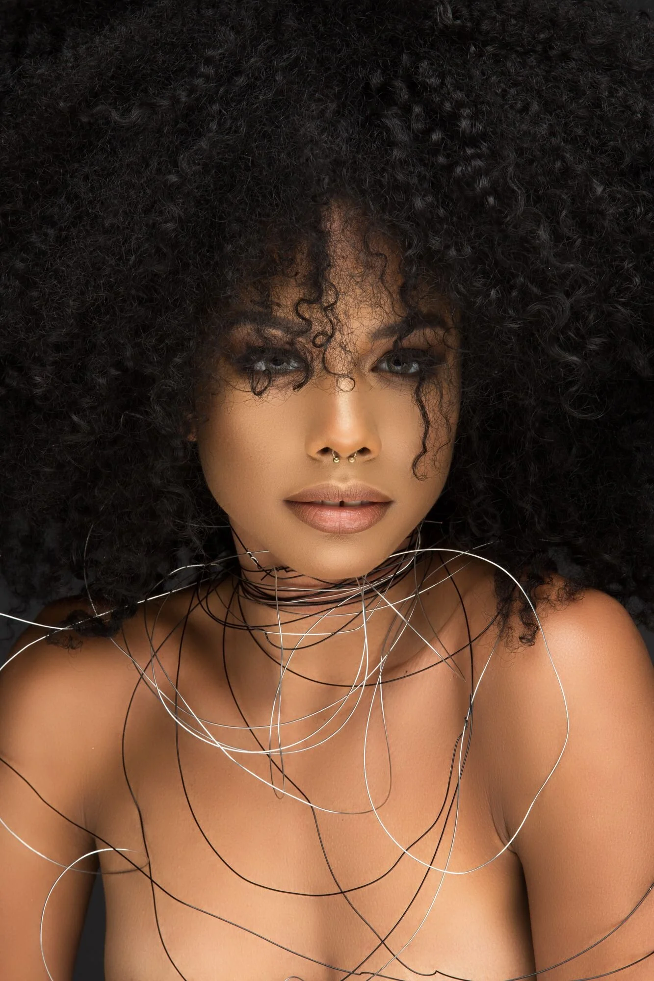 A woman with dark curly hair and light skin, wearing a metallic choker necklace and a nose ring on her septum, looking directly at the camera.