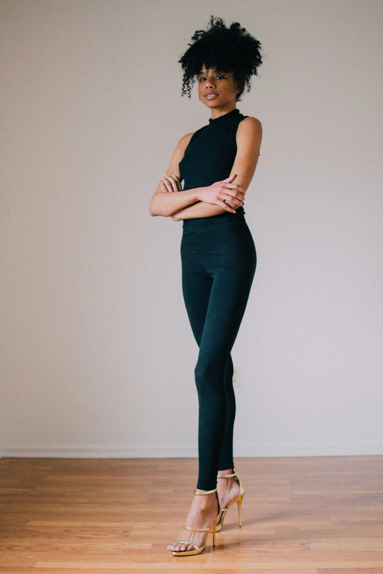 A woman standing with arms crossed, wearing a black sleeveless top, black pants, and beige high-heeled sandals, against a plain off-white wall and wooden floor.