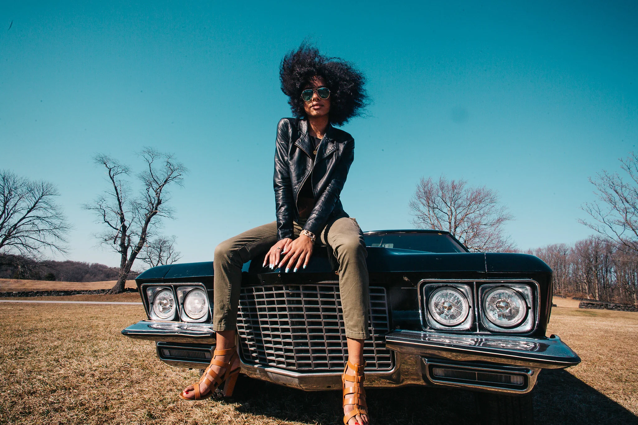 A woman with curly hair wearing sunglasses, a black leather jacket, and tan high-heeled sandals sitting on the hood of a vintage black car outdoors on a clear day with leafless trees in the background.