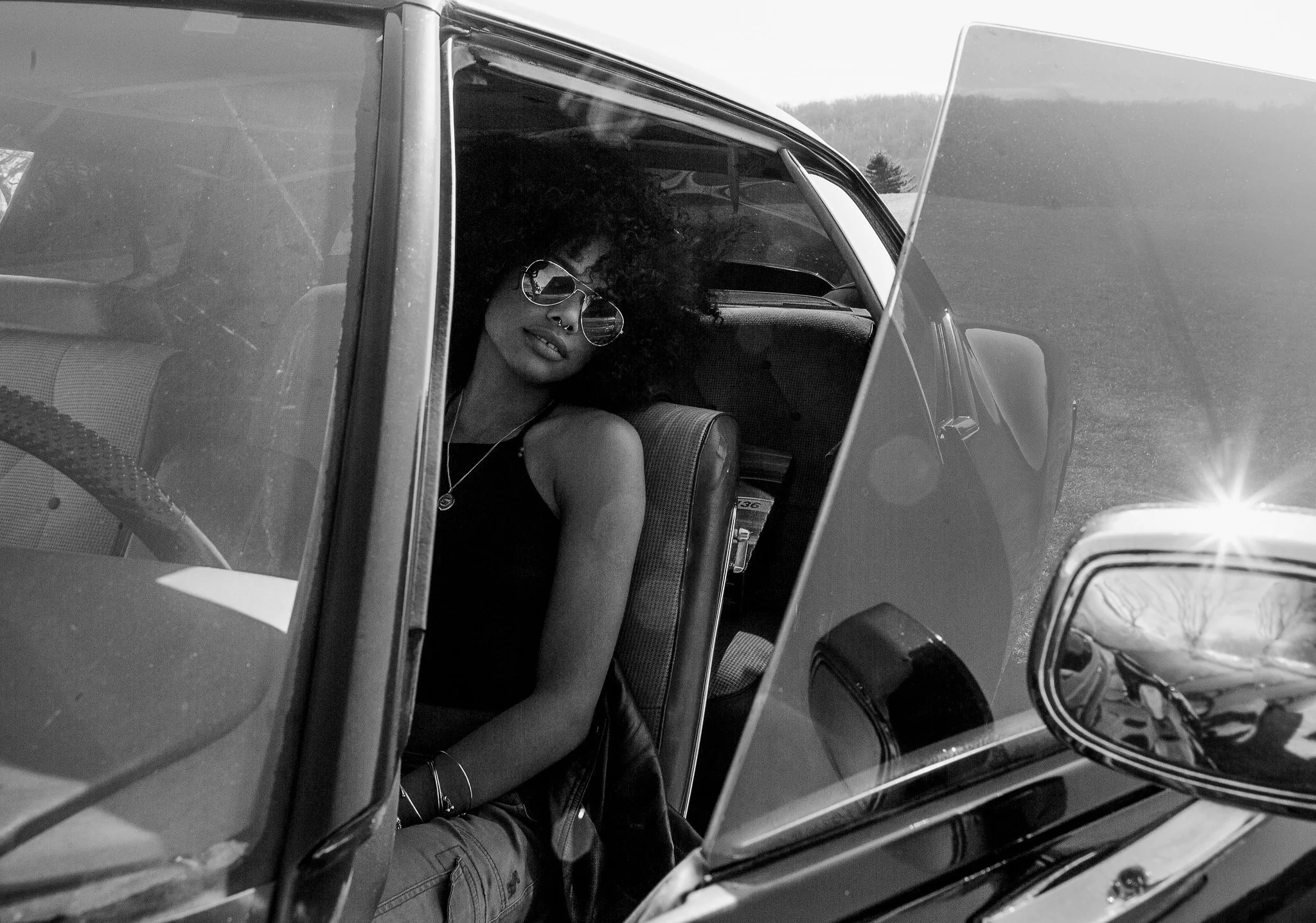 Person with curly hair and sunglasses sitting in the backseat of a car, viewed through the open window.